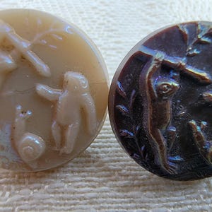 May include: Two antique buttons with relief carvings. One button is beige with a scene of a rabbit, a person, and a snail. The other button is dark brown with a monkey, a person, and a snail. Both buttons are round.