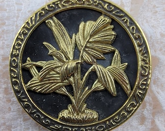 May include: A gold-coloured metal pin with a black background featuring a stylised floral design. The pin has a circular shape with a decorative border.