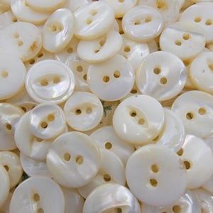 May include: A collection of off-white, pearlescent buttons with two or four holes. The buttons vary in size and shape, including round and oval designs. The buttons have a glossy finish and are scattered across the image.