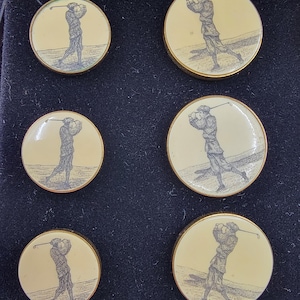 Puede incluir: Conjunto de seis botones con temática de golf. Cada botón presenta un fondo crema con una ilustración en blanco y negro de un golfista en acción. Los botones tienen un borde metálico dorado y están dispuestos en una caja negra.