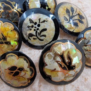 May include: A selection of vintage black buttons with iridescent shell inlays. Each button showcases a unique design, including floral and bird motifs, with gold trim. The buttons are displayed on a soft, textured surface.