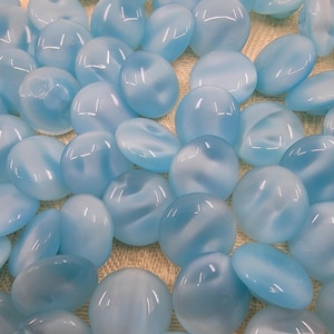 Puede incluir: Una colección de cuentas de vidrio ovaladas de color azul claro con un patrón de remolino blanco lechoso. Las cuentas varían en tamaño y forma, con un acabado liso y brillante. Las cuentas están dispersas sobre una superficie de tono neutro.
