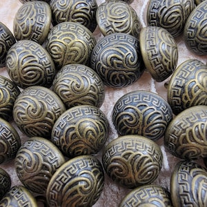May include: A collection of antique brass-colored buttons. Each button has a rounded, oval shape with a detailed, embossed design featuring a Greek key pattern and swirling motifs. The buttons are clustered together, showcasing their intricate details.
