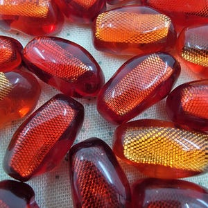 Puede incluir: Una colección de cuentas de vidrio rojas translúcidas de forma ovalada. Cada cuenta tiene un patrón texturizado en forma de rejilla en el interior, algunas aparecen más anaranjadas. Las cuentas están dispersas, creando una disposición visualmente atractiva.