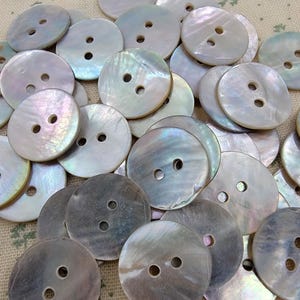 May include: A collection of iridescent, round buttons with two holes each. The buttons have a pearlescent sheen, displaying a range of colors including white, gray, and hints of pink and blue. The buttons are scattered across a textured, light-colored surface.