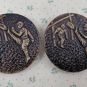 May include: Two round, brown, textured buttons with a raised design of three figures playing a game of rugby. The figures are in a light brown color.
