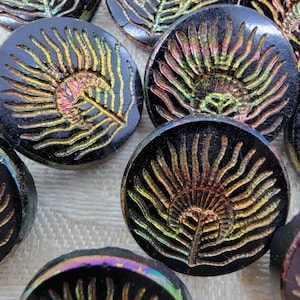 Puede incluir: Una colección de botones de vidrio negro iridiscente con un diseño de pluma de pavo real. Los botones tienen un fondo oscuro, casi negro, con un diseño de pluma de pavo real dorado e iridiscente.