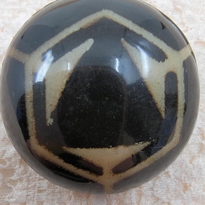 May include: A round, black decorative object with a geometric design in off-white. The design features a series of connected shapes, creating a pattern across the surface. The object has a glossy finish and appears to be made of ceramic or a similar material.