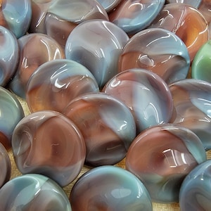 Op de afbeelding: Een verzameling gladde, ovale glazen stenen in tinten bruin, blauw en grijs. De stenen hebben een glanzend, gepolijst uiterlijk, met wervelende kleurpatronen. Het algehele effect is een natuurlijke, organische esthetiek, geschikt voor decoratieve doeleinden.