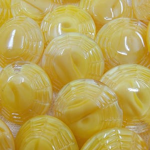 Może przedstawiać: Zbliżenie na wiele żółtych, kopulastych szklanych guzików. Każdy guzik ma teksturowany, przypominający pajęczynę wzór na powierzchni. Guziki mają błyszczące wykończenie i marmurkowy wygląd, z wirami jaśniejszego żółtego wewnątrz.