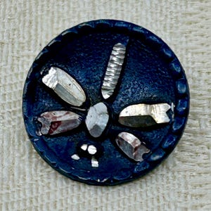 Può includere: Un bottone rotondo blu scuro con un design a libellula. Il bottone presenta accenti sfaccettati color argento disposti a forma di libellula. Il bordo del bottone ha un bordo smerlato strutturato. Il corpo della libellula è un accento argentato a coste.