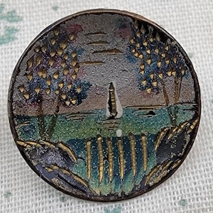 Pode incluir: Um botão redondo e antigo com uma cena de paisagem marinha pintada. O botão apresenta um veleiro na água, com árvores de cada lado. A paleta de cores inclui tons de azul, verde, dourado e roxo.