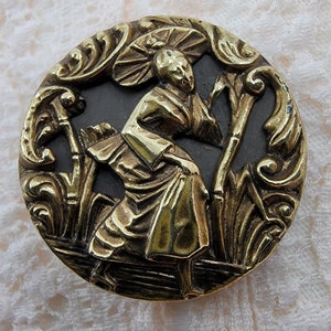 May include: A gold-toned metal button featuring a raised design of a person in a traditional Japanese outfit holding a fan. The button is set against a black background with ornate floral details.