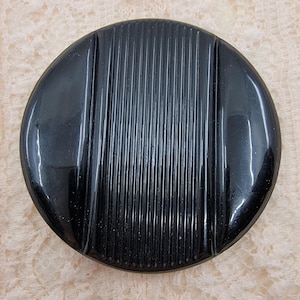 Puede incluir: Un botón redondo negro vintage con un acabado brillante. El botón presenta una serie de ranuras verticales y paralelas en su superficie, creando un diseño texturizado. La forma del botón es un círculo perfecto.