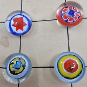 May include: Four colorful glass game pieces with intricate designs. One piece features a red star in a blue circle, another has a red and blue floral pattern, and the other two have yellow, blue, and red designs. The pieces are on a light brown surface.