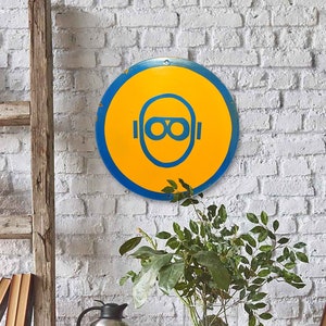 May include: A round yellow and blue sign with a graphic of goggles hangs on a white brick wall. A wooden ladder with books leans against the wall. A red table holds an orange chair, plants, and a coffee pot.
