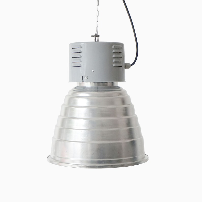Puede incluir: Una gran l&aacute;mpara colgante de estilo industrial. La l&aacute;mpara tiene una pantalla plateada escalonada y una carcasa superior gris con ranuras de ventilaci&oacute;n. Cuelga de una cadena y tiene un cable de alimentaci&oacute;n negro.