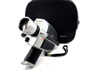 May include: A vintage Bauer CTM Super 8 movie camera with a silver and black body, lens, and handle. A black leather case is behind the camera. The camera has a wrist strap attached.