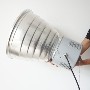 Puede incluir: Una gran l&aacute;mpara industrial de metal plateado con un reflector acanalado en forma de cono. La l&aacute;mpara tiene una carcasa rectangular gris y un cable de alimentaci&oacute;n negro. Una cadena est&aacute; unida a la carcasa.