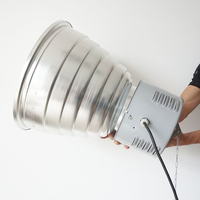 Puede incluir: Una gran l&aacute;mpara industrial de metal plateado con un reflector acanalado en forma de cono. La l&aacute;mpara tiene una carcasa rectangular gris y un cable de alimentaci&oacute;n negro. Una cadena est&aacute; unida a la carcasa.