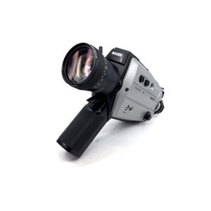 Op de afbeelding: Een vintage Bauer Super 8 filmcamera, met een grote lens en een zwarte grip met textuur. De camerabody is zilver en zwart, met de merknaam Bauer zichtbaar. De lens is zwart en heeft meetmarkeringen.