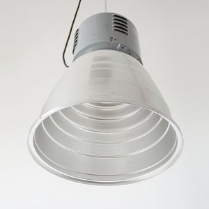 Puede incluir: Una l&aacute;mpara industrial colgante con una parte superior gris y un gran reflector plateado en forma de cono. El reflector tiene m&uacute;ltiples anillos conc&eacute;ntricos. Un cable y una cadena negros est&aacute;n unidos para colgar.