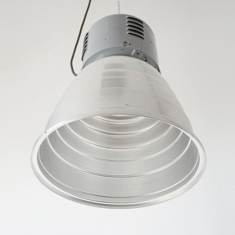 Puede incluir: Una l&aacute;mpara industrial colgante con una parte superior gris y un gran reflector plateado en forma de cono. El reflector tiene m&uacute;ltiples anillos conc&eacute;ntricos. Un cable y una cadena negros est&aacute;n unidos para colgar.