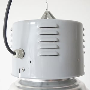 Puede incluir: L&aacute;mpara colgante industrial gris con cuerpo cil&iacute;ndrico y reflector plateado acanalado. La l&aacute;mpara tiene ranuras de ventilaci&oacute;n horizontales y un cable de alimentaci&oacute;n negro. Est&aacute; suspendida de una cadena.