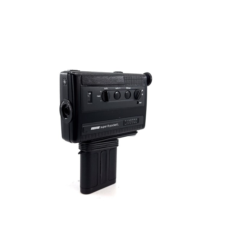 May include: A black Super 8 pocket L camera by Revue. The camera has a built-in microphone, zoom lens, and a viewfinder. The camera is mounted on a black battery pack.
