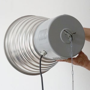 Puede incluir: Una l&aacute;mpara de estilo industrial plateada con una pantalla corrugada en forma de cono. La l&aacute;mpara tiene una carcasa gris, un cable de alimentaci&oacute;n negro y una cadena para colgar. La luz es sostenida por las manos de una persona.