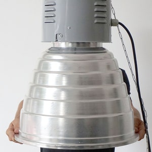 Puede incluir: Una l&aacute;mpara met&aacute;lica industrial grande con una secci&oacute;n superior gris con ranuras de ventilaci&oacute;n y un reflector plateado escalonado. La l&aacute;mpara est&aacute; suspendida por una cadena y un cable.