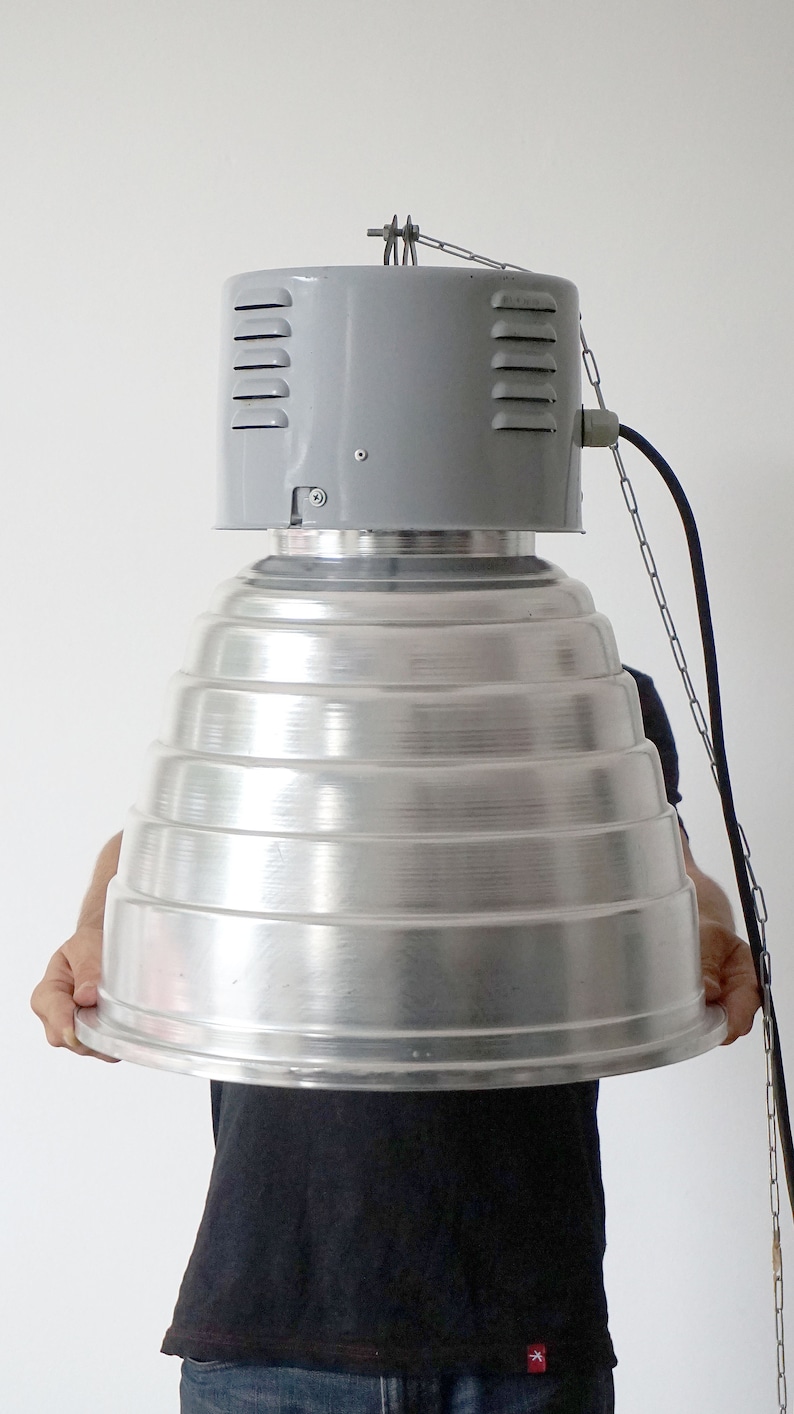 Puede incluir: Una l&aacute;mpara met&aacute;lica industrial grande con una secci&oacute;n superior gris con ranuras de ventilaci&oacute;n y un reflector plateado escalonado. La l&aacute;mpara est&aacute; suspendida por una cadena y un cable.