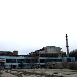 Puede incluir: Vista exterior de un edificio industrial abandonado con paredes azules y grises y numerosas ventanas. Dos altas chimeneas se alzan en el fondo contra un cielo nublado. El edificio parece desgastado y en mal estado.
