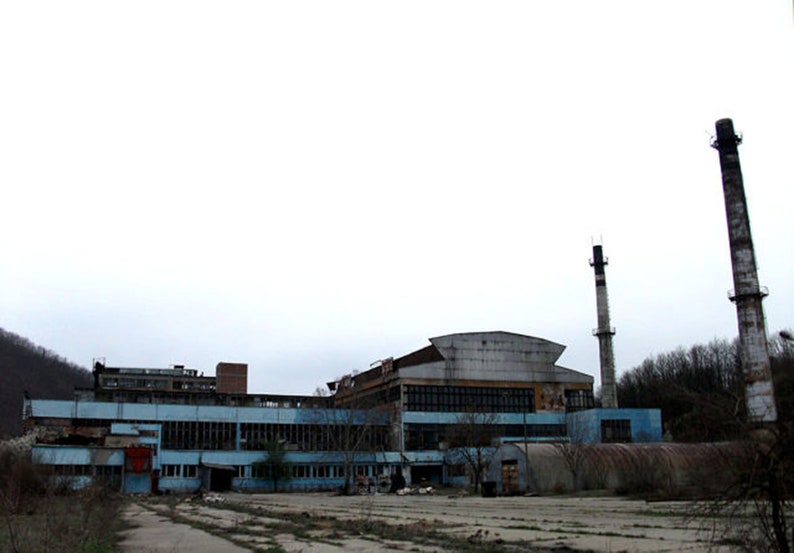 Puede incluir: Vista exterior de un edificio industrial abandonado con paredes azules y grises y numerosas ventanas. Dos altas chimeneas se alzan en el fondo contra un cielo nublado. El edificio parece desgastado y en mal estado.