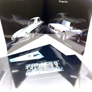 May include: A brochure for the Mercedes-Benz SL featuring a silver 1950s Mercedes-Benz SL roadster and a silver 1990s Mercedes-Benz SL roadster. The brochure text reads "Die Legende lebt" and "Der Mercedes SL" and "Mit der Technologie der Zukunft".
