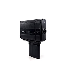 May include: A black Super 8 film camera with a textured body and a lens on the front. The camera is mounted on a black stand.