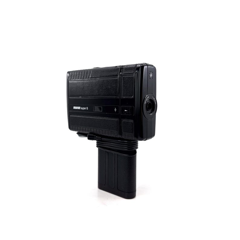 May include: A black Super 8 film camera with a textured body and a lens on the front. The camera is mounted on a black stand.