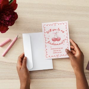 May include: A white card with pink and red text and a cherry illustration. The text reads "Will you be my bridesmaid? Having you by my side would be the cherry on top!"