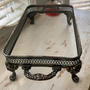 May include: An antique silver-toned rectangular serving tray with ornate detailing. The tray features decorative handles and is supported by four claw feet. The edges have a delicate, openwork design. The tray is likely made of metal.