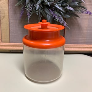 Può includere: Un barattolo di vetro trasparente con un coperchio arancione. Il barattolo ha una forma rotonda e liscia ed è perfetto per riporre piccoli oggetti.