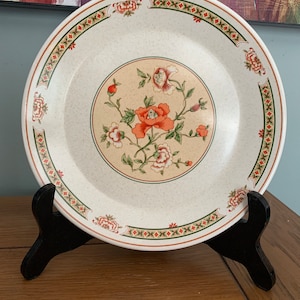 Op de afbeelding: Een wit keramiek bord met een bloemenmotief. Het bord heeft een beige achtergrond met een groene, rode en witte rand. Het midden van het bord heeft een bloemenmotief met oranje, rode en witte bloemen.
