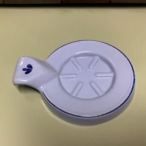 Könnte beinhalten: Weißer Keramikdeckel mit blauem Rand und Griff mit blauem Blumenmuster. Der Deckel hat eine kreisförmige Form mit einem erhabenen, sternartigen Muster in der Mitte. Ein dekorativer Küchenartikel.