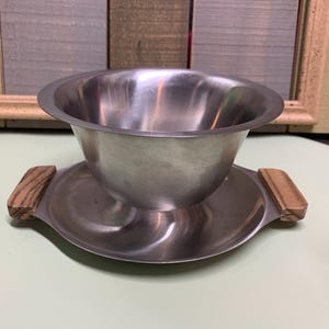 Puede incluir: Un bol para servir de acero inoxidable con asas de madera se asienta sobre un plato de acero inoxidable a juego. El bol es redondo y tiene una superficie lisa y brillante.