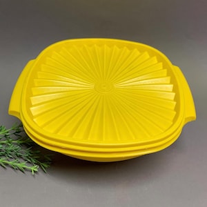 May include: Bright yellow square container with a ridged lid. The lid has a sunburst design radiating from the center. The container has rounded corners and handles on each side. The container is made of plastic.