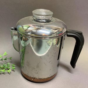May include: A vintage stainless steel coffee percolator with a clear glass lid and a black handle. The coffee pot has a shiny, reflective surface and a small spout. The bottom of the pot has a brown ring.