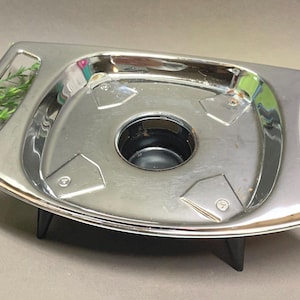 Puede incluir: Un chafing dish plateado vintage con un área central empotrada y un asa negra. El plato tiene un diseño geométrico y se asienta sobre una base negra. Es una pieza para servir para mantener la comida caliente.