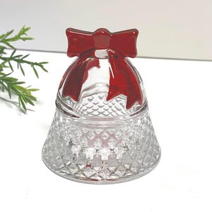 Puede incluir: Un tarro de vidrio transparente en forma de campana con un lazo rojo en la parte superior. El tarro tiene un patrón de diamantes en la mitad inferior y detalles en rojo en la mitad superior. El tarro es probablemente para fines decorativos.