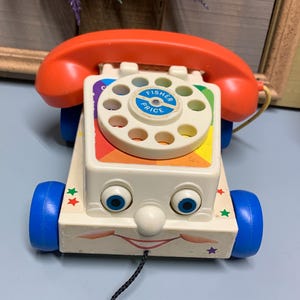 May include: A vintage Fisher-Price toy phone with a pull string, colorful buttons, and a smiling face. The phone has a red handset, blue wheels, and a white body with rainbow accents.