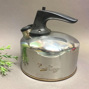 Puede incluir: Una tetera de acero inoxidable con asa y boquilla negras. La tetera tiene un cuerpo redondeado y un borde dorado. El nombre de la marca está grabado en relieve en el lateral. Un artículo de cocina clásico.