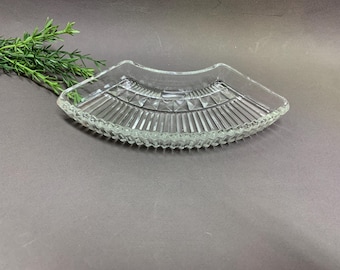 May include: A clear glass dish shaped like a fan with a scalloped edge and a textured pattern of vertical ribs and small raised diamond shapes along the top border. The dish is transparent and features a curved design with a wide, flat base.